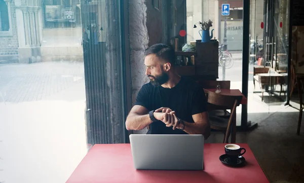 Young Bearded Businessman Wearing Black Tshirt Working Laptop Urban Cafe.Man Sitting Wood Table Cup Coffee Looking Through Window Touch Smartwatch.Coworking Process Business Startup.Color Filter.