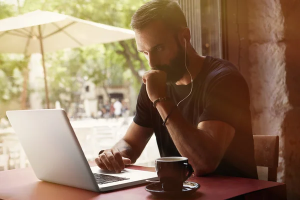 Koncentrerad ung skäggiga affärsman bär svart tshirt arbetar laptop Urban Cafe. man sittande bord kopp kaffe lyssnar musik. Coworking process Business Startup. suddig bakgrund. färg filter. — Stockfoto