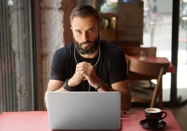 Snygg ung Bearded affärsman klädd i svart tshirt arbetar laptop Urban Cafe. man sittande träbord kopp kaffe lyssnar musik. Coworking process Business Startup. suddig bakgrund. — Stockfoto