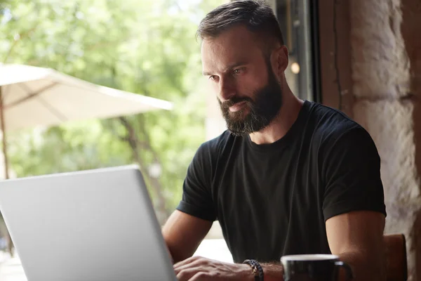 Concentrated Bearded Man Wearing Black Tshirt Working Laptop Wood Table Urban Cafe.Young Manager Work Notebook Modern Interior Design Loft Place.Coworking Process Business Startup.Color Filter. Stock Picture