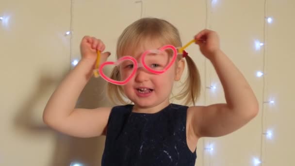Graciosa niña en gafas en forma de corazón de neón mira a la cámara. — Vídeos de Stock