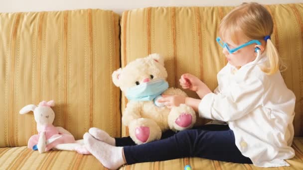 Nettes Vorschulmädchen in weißer Arztuniform und Brille behandelt Spielzeug-Teddybär mit Stethoskop — Stockvideo