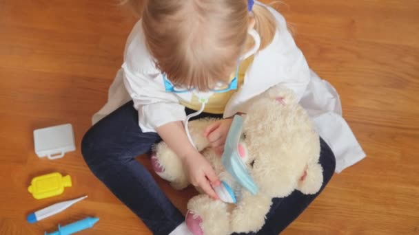 Милая дошкольница в белой медицинской форме и очках лечит игрушечного плюшевого мишку стетоскопом — стоковое видео