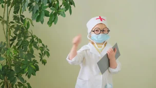 Tıp üniformalı, gözlüklü ve koruyucu maskeli sevimli küçük kız baş parmağını kaldırmış ve dans ediyor. — Stok video