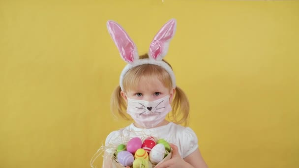 Ragazza carina con le orecchie di coniglio in una maschera medica mostra le uova di Pasqua e mostra il tuo sorriso senza maschera — Video Stock