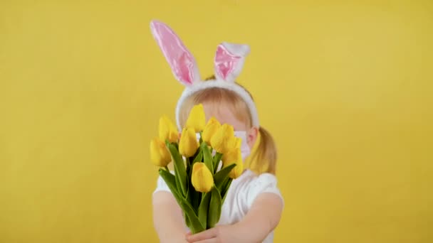 Koruyucu tıbbi maskeli sevimli tavşan kulaklı kız hediye olarak bir buket sarı bahar çiçeği tutuyor. — Stok video