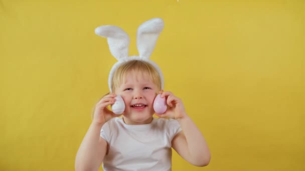 Portrét veselé blond holčička hrající si s velikonočním vejcem na žlutém pozadí. — Stock video