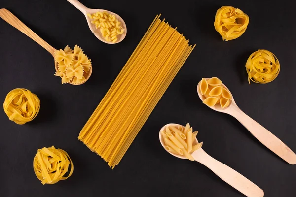 Different pasta types in wooden spoons on black table. — Stock Photo, Image