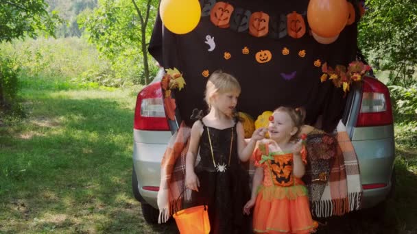 Dos hermanitas celebrando Halloween y comiendo dulces de cubos sentadas en el maletero del coche. — Vídeos de Stock