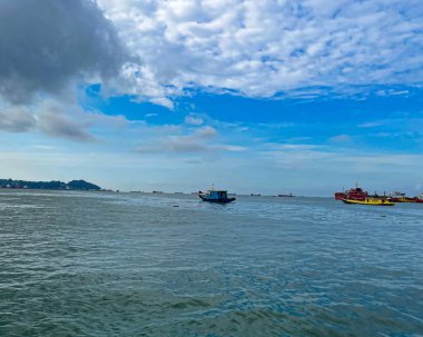 Mavi balıkçı teknesi engin açık sularda çok sayıda kargo gemisi ve bulutlu bir gökyüzünün altında puslu bir kıyı şeridiyle yol alır.