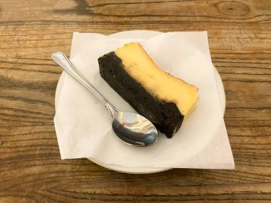 Zengin, koyu bir tabanı kremsi açık bir üstle birleştiren lezzetli bir tatlı dilimi, ahşap bir yüzeyde gümüş bir kaşıkla beyaz bir tabakta sunuluyor.