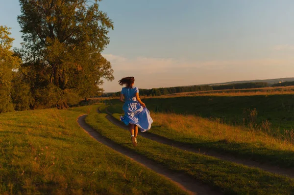 Šťastná dívka v modrých šatech se zády ke kameře, běží během západu slunce. Stock Fotografie