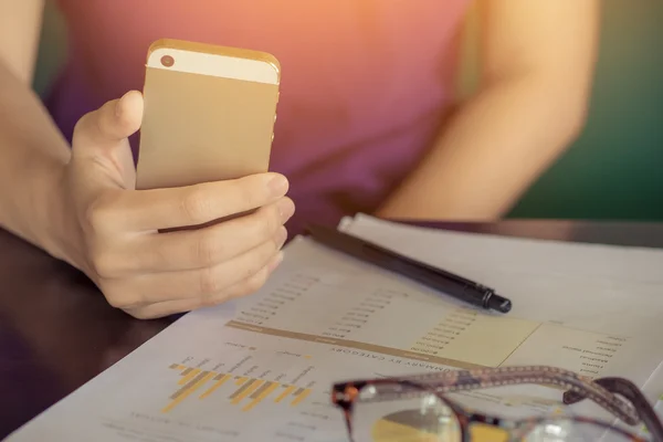 Young business woman working from home using smart phone with payoung business woman working from home using smart phone with paper chart graph and glasses on wooden desk.  Vintage filter effect. — Stok fotoğraf