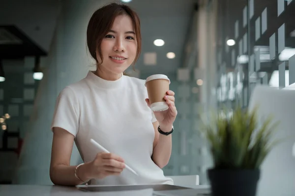 Cretive woman in white dress smiling holding stylus pencil and coffee in home office. Work from home concept. - Stock-foto