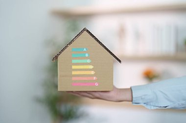 A hand holds a cardboard house model displaying energy efficiency ratings, symbolizing sustainability and eco-friendly living.