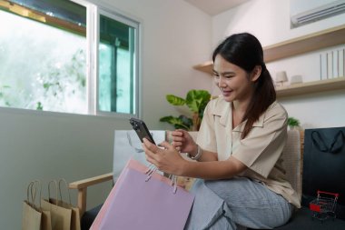 Genç bir kadın alışveriş için akıllı telefonunu kullanırken gülümsüyor. Etrafında renkli çantalar ve yanında kedisiyle..