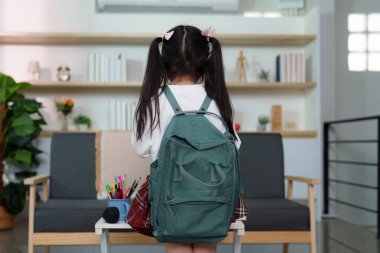 Sırt çantalı genç bir kız kameraya arkası dönük durur, şık bir ev ortamında okula hazır olmayı sembolize eder.