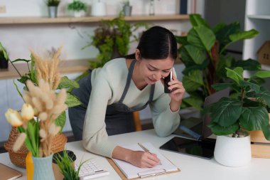 Bir kadın telefonda konuşurken, çalışma odasındaki yemyeşil bitkilerle çevrili notlar yazar..