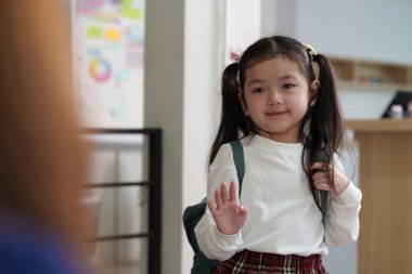 Genç bir kız, önümüzdeki okul günü için heyecanını ifade ederek annesine neşeyle el sallıyor. Sevinç ve bağla dolu bir an.