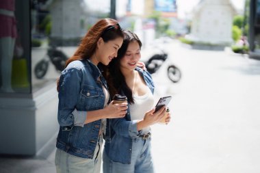 İki arkadaş gülümsüyor ve açık havada akıllı bir telefona bakarken neşeli bir anı paylaşıyor, arkadaşlığı ve mutluluğu somutlaştırıyor..