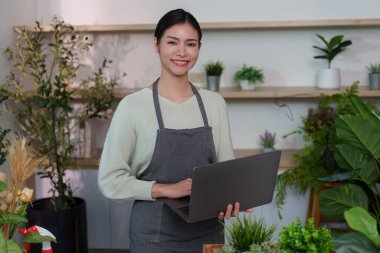 Bir kadın canlı, kapalı bir bahçede dizüstü bilgisayar kullanarak bitki işlerini gülümseyerek yürütür..