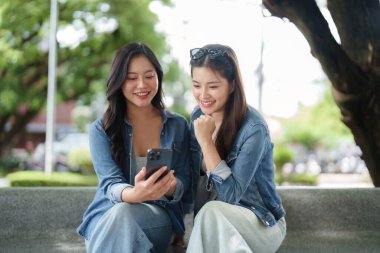 İki arkadaş birlikte bir bankta oturup gülüşüyorlar ve canlı bir şehir parkında kahvelerini yudumlarken akıllı telefonlarına bakıyorlar..