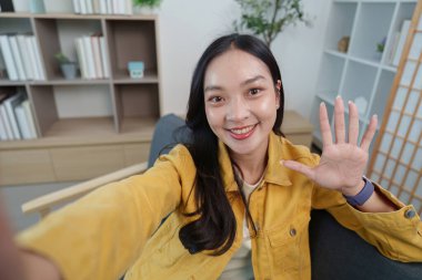 Gülümseyen bir kadın kameraya el sallıyor, şık evinde neşeli bir anı yakalıyor, canlı bir yaşam tarzını yansıtıyor..