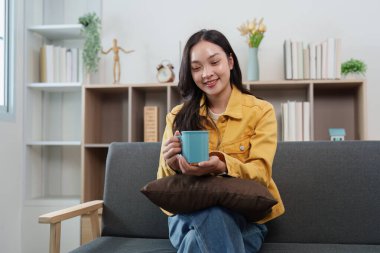 Genç bir kadın elinde kupa tutarken gülümsüyor, modern kapalı bir ortamda rahat bir yaşam tarzını temsil ediyor..