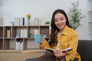 Sarı ceketli neşeli bir kadın bir kitap ve kahveyle rahatlıyor, şık evinde rahat ve neşe dolu bir yaşam tarzını somutlaştırıyor..