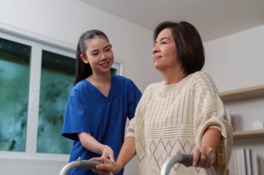 Bir hemşire yaşlı bir kadına yürüteçle yardım eder, ev ortamında dolaşmanın ve bakımın önemini vurgular..