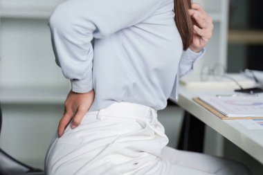 Bir kadın masasında otururken belini rahatsızlık içinde tutuyor ve sabit çalışmanın sağlık üzerindeki etkisini vurguluyor..