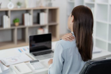 Genç bir kadın masasında omzuna masaj yapıyor ve ofis sendromunun sağlığına olan fiziksel etkisini gösteriyor..