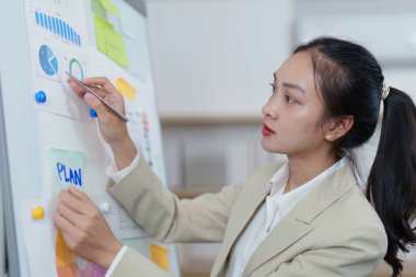 Kararlı bir kadın çizelgeleri ve notları beyaz tahtada inceler. Profesyonel bir ortamda analitik yetenekler sergiler..