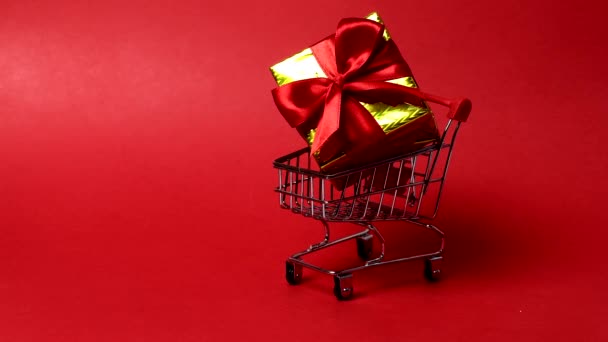 Vozík v supermarketu hraček na červeném pozadí. Vozík má dárkovou krabici a vánoční míčky. Novoroční nákupní koncept. Stříbrná koule. — Stock video