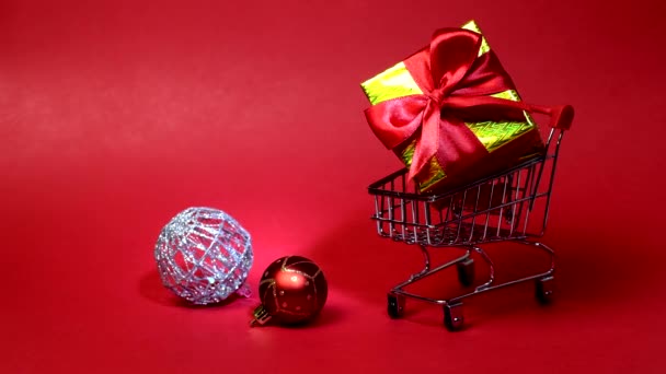 Carro de supermercado de juguete sobre fondo rojo. El carro tiene caja de regalo y bolas de Navidad. Concepto de compras de Año Nuevo. Luz en el carro. — Vídeo de stock