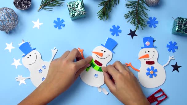 Uma criança está fazendo um presente de Natal com um boneco de neve. Árvore de Natal ornamentos pendurados. Peças de boneco de neve em fundo de madeira azul. Natal artesanato ideias. — Vídeo de Stock