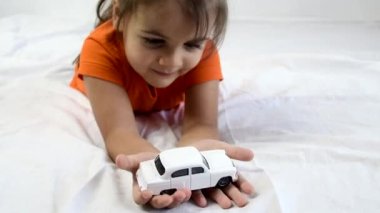 Küçük kız beyaz oyuncak arabayla oynuyor. Beyaz çarşafın üzerinde yatıyordu. Gülümsüyor.