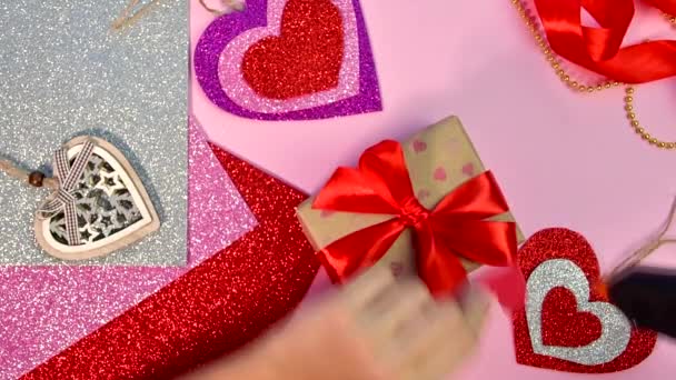 Valentinsdag eller fødselsdagsgaver. Valentinsdag gaver med et rødt papir hjerte på et træbord. Pige gør rødt hjerte fra rødt papir. Lyserød baggrund. Set fra oven. – Stock-video
