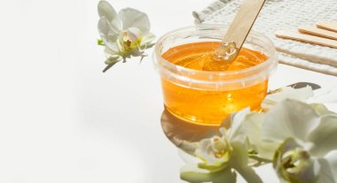 Şeffaf bir kavanozda bal ya da balmumu ezmesi ve beyaz arka planda beyaz orkide. Şekerleme. Deplasyon ve güzellik konsepti. Ağda yapıyorum. Yumuşak odak.