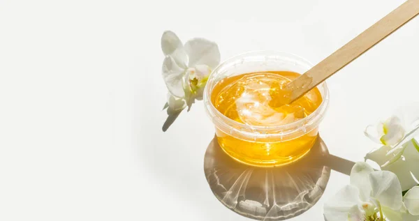 Şeffaf bir kavanozda bal ya da balmumu ezmesi ve beyaz arka planda beyaz orkide. Şekerleme. Deplasyon ve güzellik konsepti. Ağda yapıyorum. Pankart