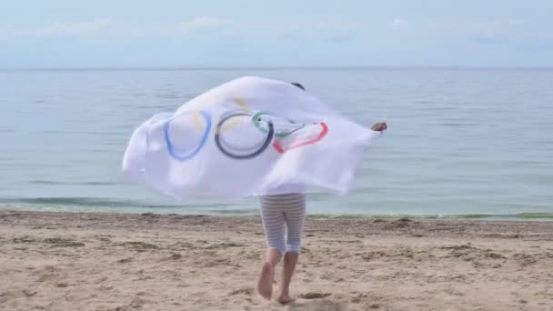 Mädchen schwenken Flagge der Olympischen Spiele im Freien über bewölkten Himmel und blauem Wasser des Meeres. Kindersportfan. 27.06.2020 St.Petersburg Russland — Stockvideo