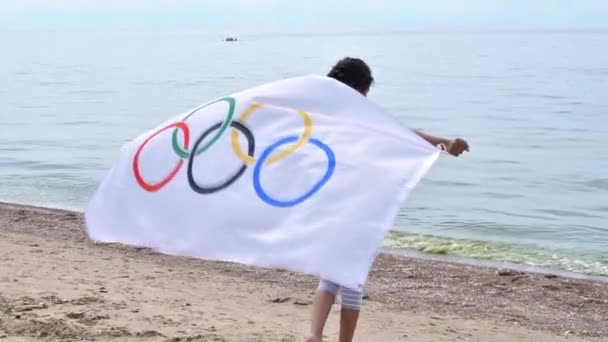 Chica ondeando bandera de los Juegos Olímpicos al aire libre sobre el cielo nublado y el agua azul del mar. Niños fanáticos de los deportes. 27.06.2020 San Petersburgo Rusia — Vídeo de stock