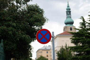 Bir Avrupa şehrinde, tarihi binalar ve yeşil ağaçların önünde park ve durmanın (durmak / durmak yok) yasak olduğunu gösteren bir trafik işareti.