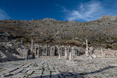 Antik Roma çeşmesi (Antoninus Fountain) ve arka planda engebeli bir dağ manzarası olan Agora 'nın taş döşeli zemini. Tarih ve mimari.