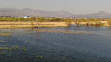 Uçsuz bucaksız bir sulak arazide süzülen insansız hava aracı görüntüleri; arka planda görkemli dağlar, yerleşimler ve endüstriyel tesisleri olan huzurlu bir doğal manzara..