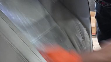 Turuncu eldiven giyen bir profesyonel temizlik köpüğü sürdükten sonra lekeleri çıkarmak için araba koltuğunu süngerle fırçalıyor. Detaylı iç temizlik aşaması.