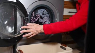 Ev ve günlük hayat temalı video klipte kırmızı ceketli bir adam çamaşır makinesinden temiz giysileri çıkarıp bir sepeti dolduruyor..