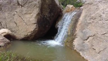 Beautiful small waterfall flowing between large rocks and green grass in a peaceful mountain landscape, offering a serene and natural scene.