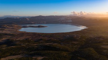 Altın saat boyunca dağların arasındaki göl havadan görülür. Uzak kar tepeleri ve gün batımı bulutlarıyla panoramik bir manzara..