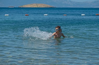 Yüzme gözlüğü takan, elleriyle su sıçratan ve derin mavi denizde eğlenen bir çocuğun fotoğrafı..